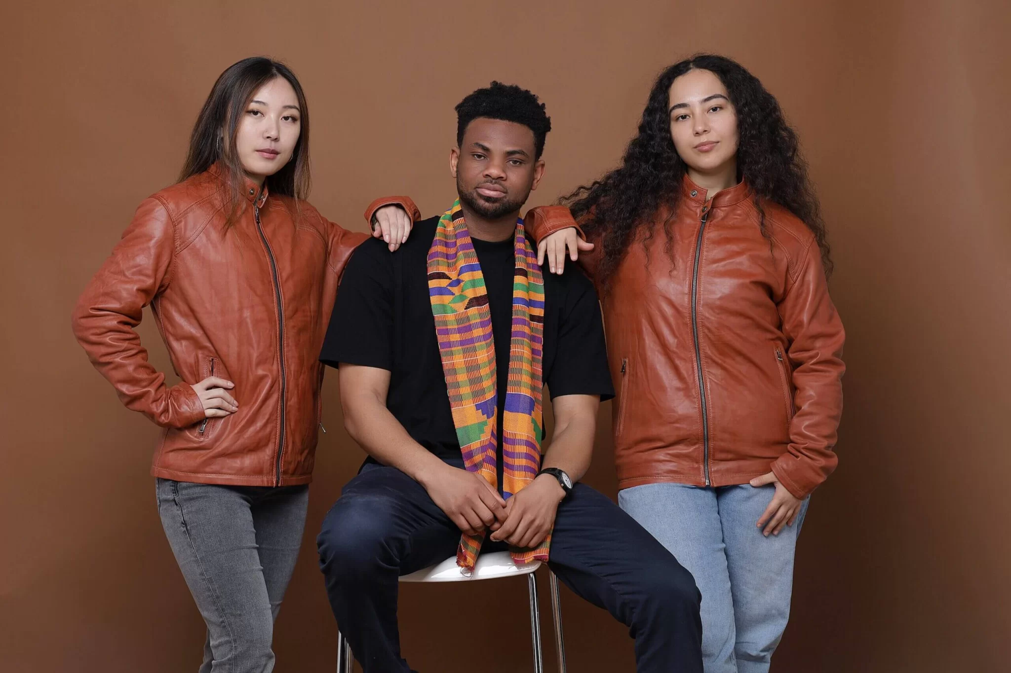 Three young creatives posing confidently, with two wearing brown leather jackets and one wearing a traditional Kente cloth scarf, representing culture and style in Nottingham.