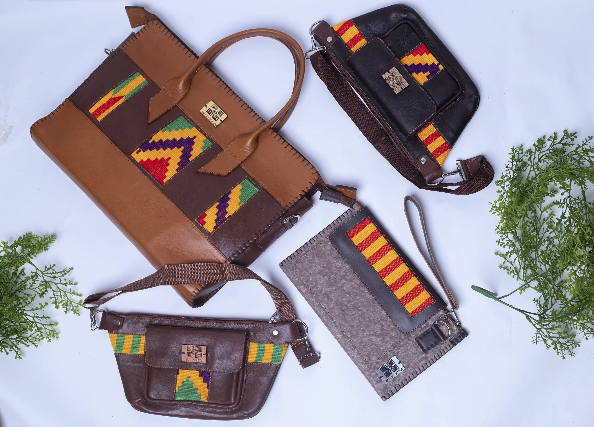 A collection of four stylish Ghanaian Kente handbags and pouches arranged on a light background with green foliage accents. The items include a brown leather satchel with vibrant Kente patterns in green, yellow, and red, a black leather satchel with multicolored Kente designs, a brown leather waist bag with colorful Kente accents, and a gray leather pouch with bold red and yellow Kente stripes, showcasing cultural craftsmanship and fashion.
