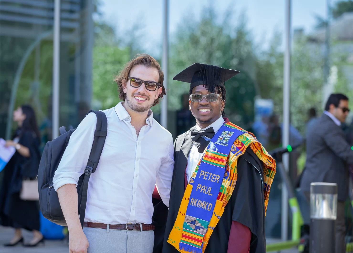 Two people standing and smiling. One person is wearing a black graduation gown, mortarboard, and a multicolored stole that has the name "PETER PAUL AKANKO" on it. The other person is wearing a white button-down shirt and sunglasses.