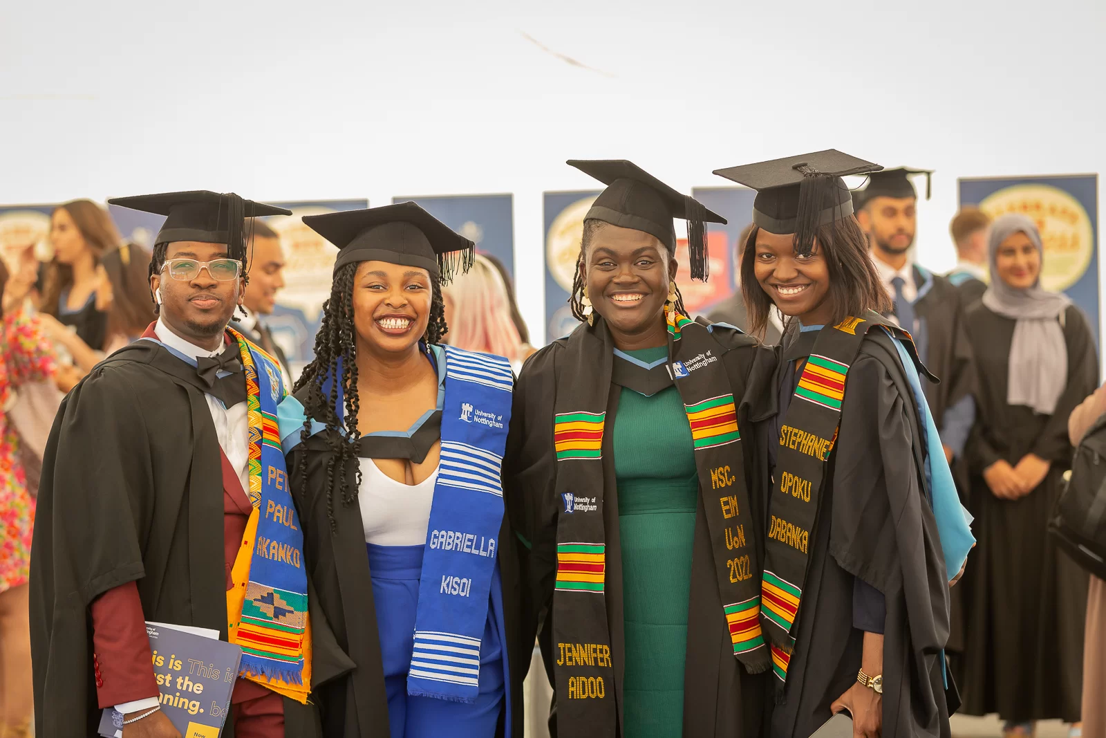 Four smiling graduates in black gowns and mortarboards, standing together for a photo. The graduates are wearing colorful stoles with embroidered names. The stoles feature a variety of patterns, including blue and white horizontal stripes, and red, yellow, and green stripes.
